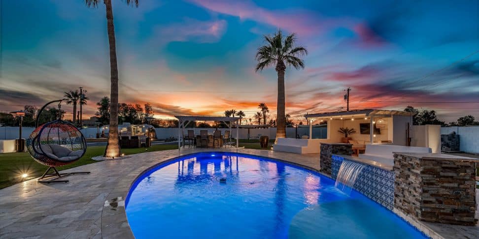 A backyard with a pool, palm trees, and a small waterfall at sunset.