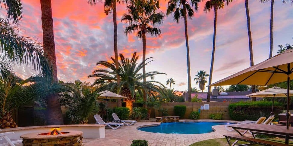 A backyard of a vacation rental in Arizona with palm trees, a pool, and a fireplace at sunset.
