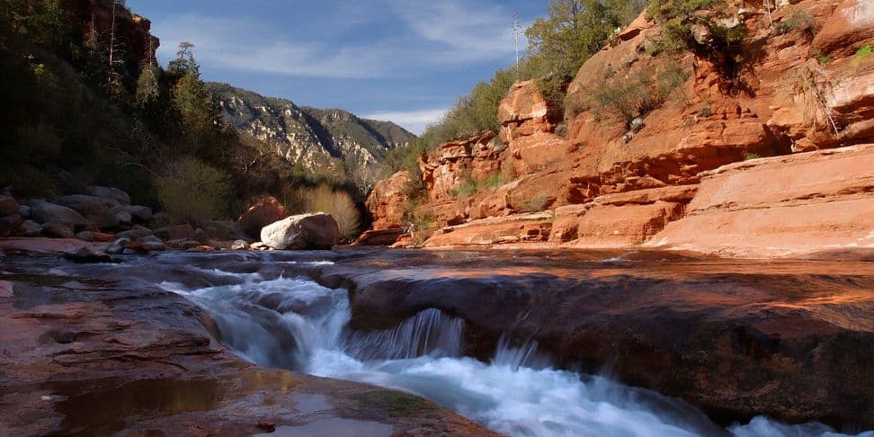 Slide Rock State Park, with a natural water slide carved into the red sandstone. Clear water rushes over smooth, terraced rocks, nestled in a canyon surrounded by towering cliffs and a forested mountain backdrop.