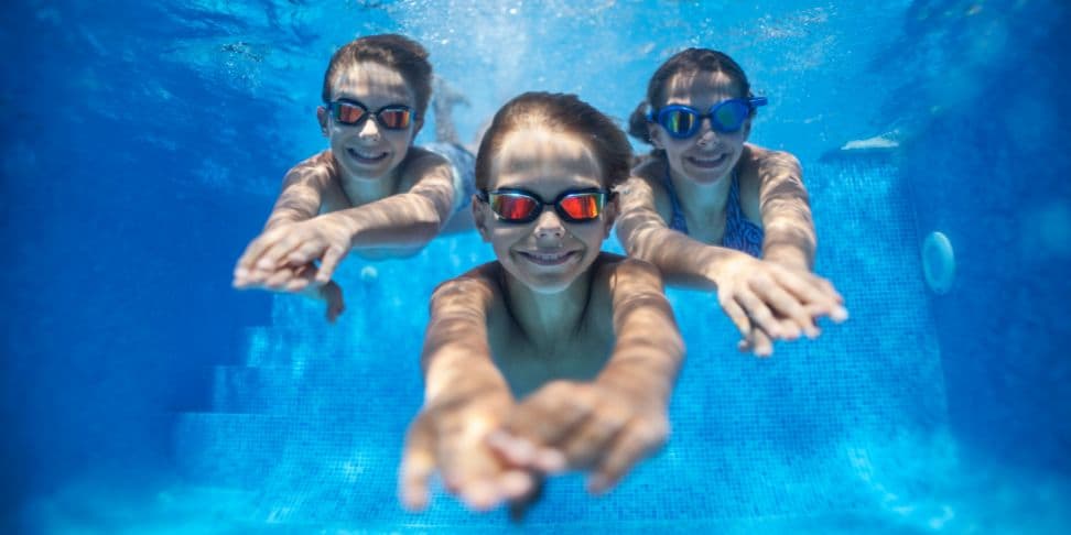 Three children swimming underwater in a clear blue pool, reaching out towards the camera with smiles visible through their goggles.