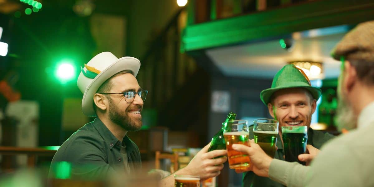 A group of friends celebrating St. Patrick's Day in a pub, with one man in the foreground wearing a white hat with an orange and green feather and glasses, smiling broadly, and toasting with a pint of beer. Another man, wearing a green leprechaun hat, smiles and clinks his beer glass in return. They are surrounded by a festive atmosphere with green lights illuminating the background.