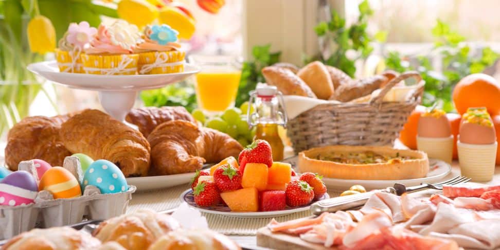 An inviting Easter brunch table is set with a variety of foods in a bright, sunlit room. In the foreground, there are colorful painted Easter eggs in a carton, a plate of fresh strawberries and cantaloupe, and flaky croissants. The middle of the table features a basket filled with an assortment of bread, while the background showcases a cake stand with decorated Easter-themed cupcakes, and a tart. The scene is completed with servings of sliced meats, glasses of orange juice, and additional Easter eggs, creating a festive and abundant holiday meal.