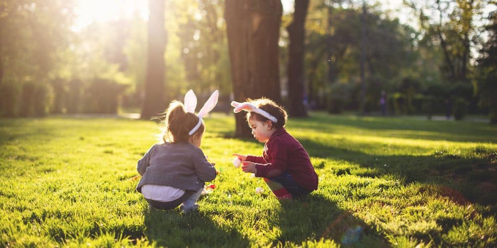Two young children with bunny ears are sitting on a lush green lawn, engrossed in play with colorful Easter eggs. 