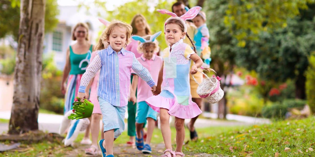 A group of children wearing pastel-colored clothes and bunny ears headbands are holding hands and happily walking down a sidewalk during an Easter event.