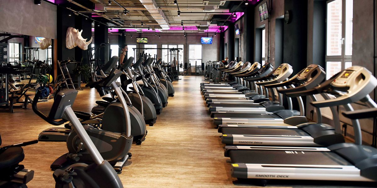 A modern gym with a row of treadmills in the foreground, positioned to face a window. Each treadmill has a digital display. In the background, other gym equipment like stationary bikes and weights are visible. The space is well-lit with a combination of natural light from the windows and overhead lights.
