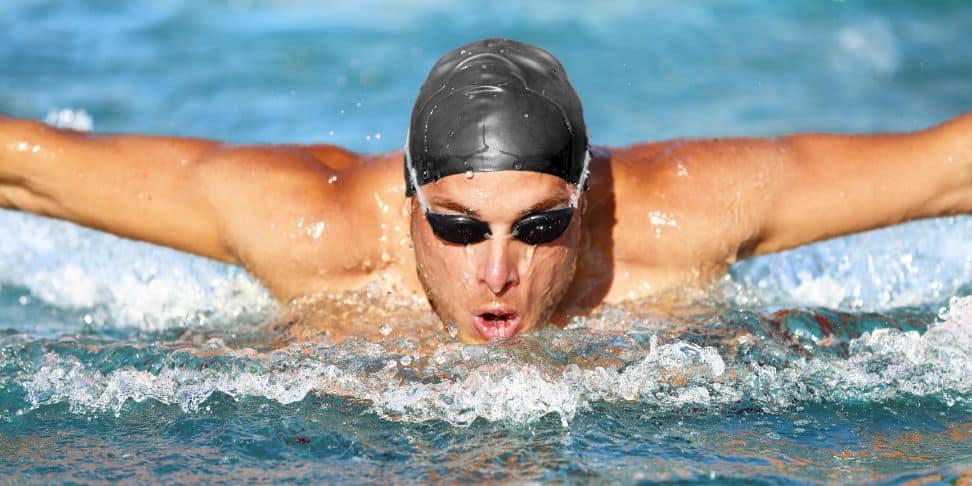 A close-up action shot of a swimmer doing the butterfly stroke in a pool. The swimmer, wearing a black swim cap and goggles, is captured with arms extended mid-stroke, creating splashes of water around. The swimmer's face is concentrated and slightly above the water's surface, exhaling.