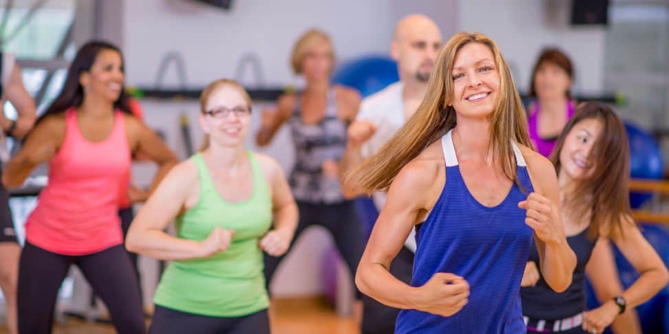 A group of smiling people engaged in a dance fitness class indoors. The focus is on a woman with long hair in the foreground, wearing a blue tank top. She looks happy and energetic. In the background, classmates of diverse ages and ethnicities are also visible, some blurred in motion, wearing brightly colored workout attire.