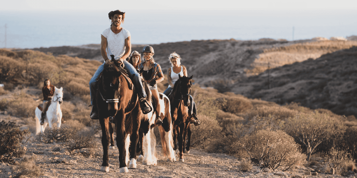 A group of people enjoying a horseback ride in a desert-like landscape during the late afternoon, with the setting sun casting a warm glow on the scene.