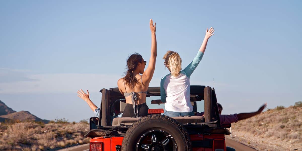 People riding in the back of a red Jeep with their hands raised on a desert road.